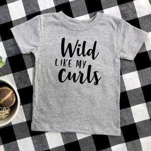 May include: A gray short-sleeved t-shirt with the text "Wild like my curls" printed in black.