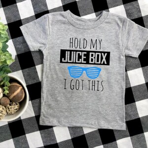 Op de afbeelding: Een grijs T-shirt met korte mouwen met de tekst "HOLD MY JUICE BOX I GOT THIS" in zwart en blauw. Het shirt heeft een blauwe zonnebrilafbeelding. Het shirt wordt weergegeven op een zwart-wit geruit oppervlak.