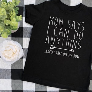 May include: Black t-shirt with white text that reads "Mom says I can do anything... except take off my bow."  The text is accompanied by a small arrow.