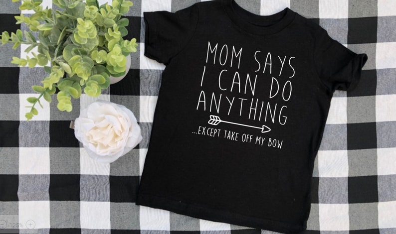 May include: Black t-shirt with white text that reads "Mom says I can do anything... except take off my bow."  The text is accompanied by a small arrow.