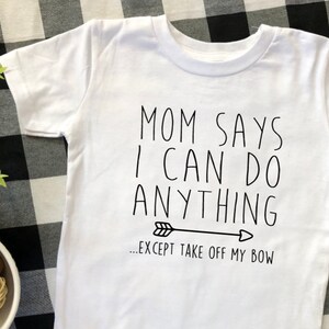 May include: White t-shirt with black text that reads "Mom says I can do anything...except take off my bow" with a black arrow pointing to the right.