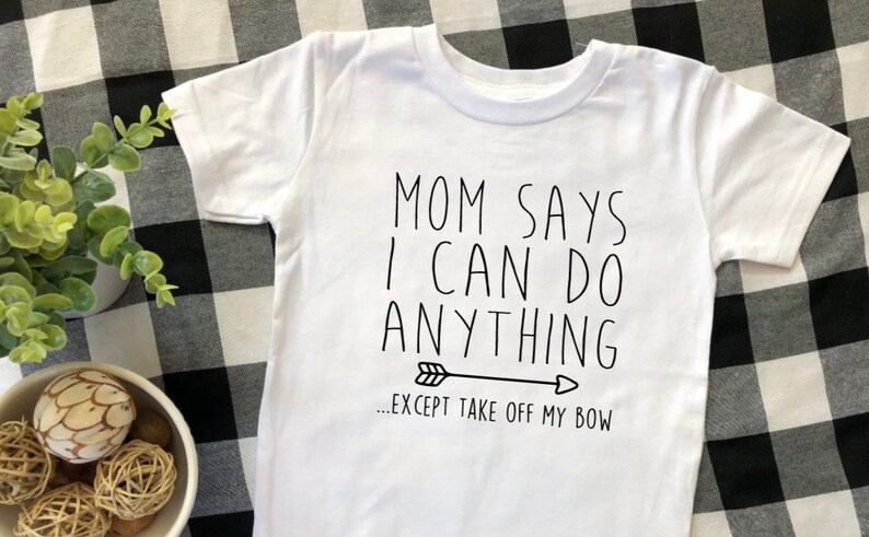 May include: White t-shirt with black text that reads "Mom says I can do anything...except take off my bow" with a black arrow pointing to the right.