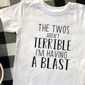 May include: White t-shirt with black text that reads "The Twos Aren't Terrible I'm Having A Blast".