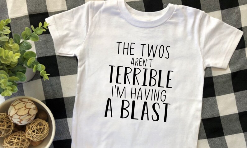 May include: White t-shirt with black text that reads "The Twos Aren't Terrible I'm Having A Blast".