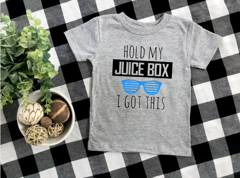 Op de afbeelding: Een grijs T-shirt met korte mouwen met de tekst "HOLD MY JUICE BOX I GOT THIS" in zwart en blauw. Het shirt heeft een blauwe zonnebrilafbeelding. Het shirt wordt weergegeven op een zwart-wit geruit oppervlak.