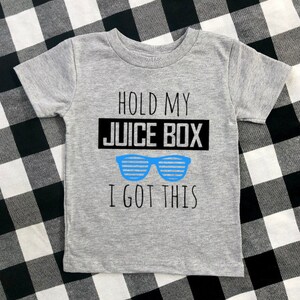 Op de afbeelding: Een grijs T-shirt met korte mouwen met de tekst "HOLD MY JUICE BOX I GOT THIS" in zwart en een blauwe zonnebril. Het shirt wordt gepresenteerd op een zwart-wit geruit oppervlak.