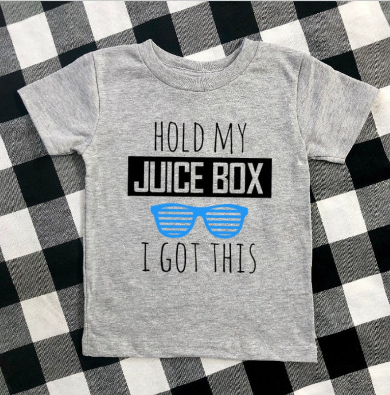 Op de afbeelding: Een grijs T-shirt met korte mouwen met de tekst "HOLD MY JUICE BOX I GOT THIS" in zwart en een blauwe zonnebril. Het shirt wordt gepresenteerd op een zwart-wit geruit oppervlak.
