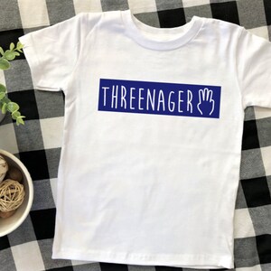 May include: White t-shirt with a blue rectangle that says "THREEENAGER" with a hand drawn three fingered hand.