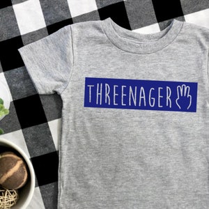 May include: A grey t-shirt with a blue rectangle that says "THREE-NAGER" with a hand with three fingers raised.