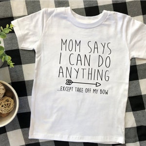 May include: White t-shirt with black text that reads "Mom says I can do anything... except take off my bow" with a black arrow pointing to the right.