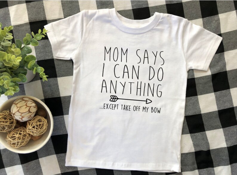 May include: White t-shirt with black text that reads "Mom says I can do anything... except take off my bow" with a black arrow pointing to the right.