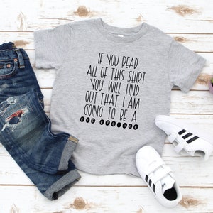 May include: A gray t-shirt with the text "IF YOU READ ALL OF THIS SHIRT YOU WILL FIND OUT THAT I AM GOING TO BE A BIG BROTHER" printed in black.