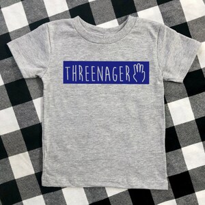 May include: Grey short-sleeved t-shirt with a blue rectangle that says "THREEENAGER" with a handprint icon.