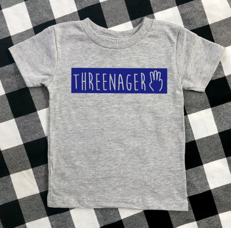 May include: Grey short-sleeved t-shirt with a blue rectangle that says "THREEENAGER" with a handprint icon.