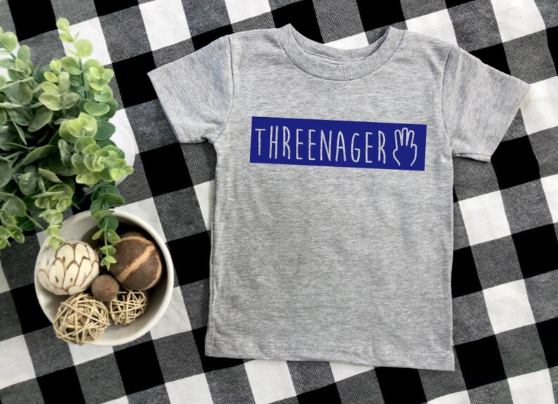 May include: Grey short-sleeved t-shirt with a blue rectangle that says "THREEENAGER" with a hand graphic.