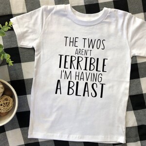 May include: White t-shirt with black text that reads "The Twos Aren't Terrible I'm Having A Blast".