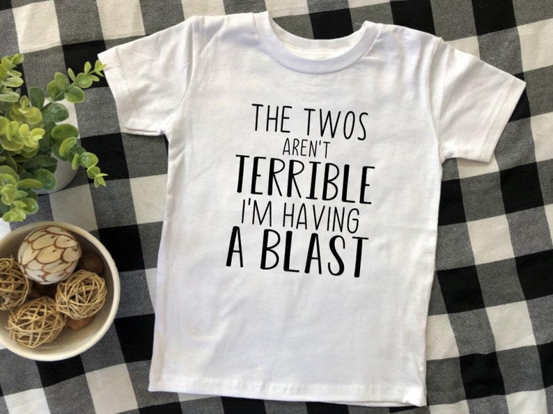 May include: White t-shirt with black text that reads "The Twos Aren't Terrible I'm Having A Blast".