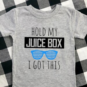 Op de afbeelding: Een grijs T-shirt met korte mouwen met de tekst "HOLD MY JUICE BOX I GOT THIS" in het zwart en een blauwe zonnebril. Het shirt is gemaakt van een zacht materiaal.