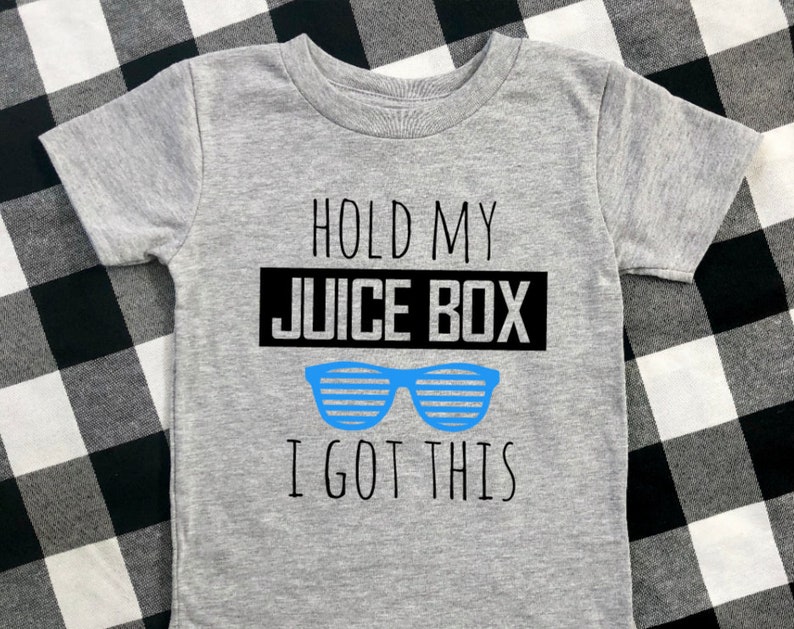 Op de afbeelding: Een grijs T-shirt met korte mouwen met de tekst "HOLD MY JUICE BOX I GOT THIS" in het zwart en een blauwe zonnebril. Het shirt is gemaakt van een zacht materiaal.