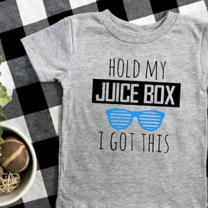 Op de afbeelding: Een grijs t-shirt met korte mouwen met de tekst "HOLD MY JUICE BOX I GOT THIS" in zwart en blauw. Het shirt heeft een blauwe zonnebrilafbeelding. Het shirt wordt gepresenteerd op een zwart-wit geruit oppervlak.