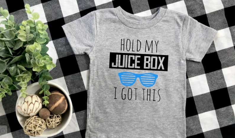 Op de afbeelding: Een grijs t-shirt met korte mouwen met de tekst "HOLD MY JUICE BOX I GOT THIS" in zwart en blauw. Het shirt heeft een blauwe zonnebrilafbeelding. Het shirt wordt gepresenteerd op een zwart-wit geruit oppervlak.