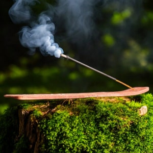 May include: A wooden incense holder with a brown incense stick burning and releasing white smoke. The holder is placed on a mossy green tree stump.
