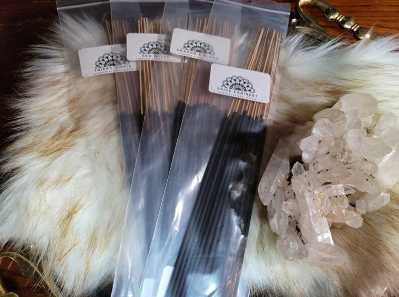 May include: Four packs of incense sticks in clear plastic packaging. The incense sticks are black and have a gold-colored string attached to the top. The packaging has labels with the names of the scents: "Trippy Hippie", "Sea Witch", "Dragon's Blood", and "Spice Cabinet".