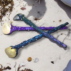 May include: Two iridescent spoons with seashell bowls and pearl accents. The spoons are crossed and lying on a white sandy surface.