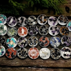 May include: Twenty-four small round containers filled with various colorful stones and crystals. Each container is sealed with a clear resin top.