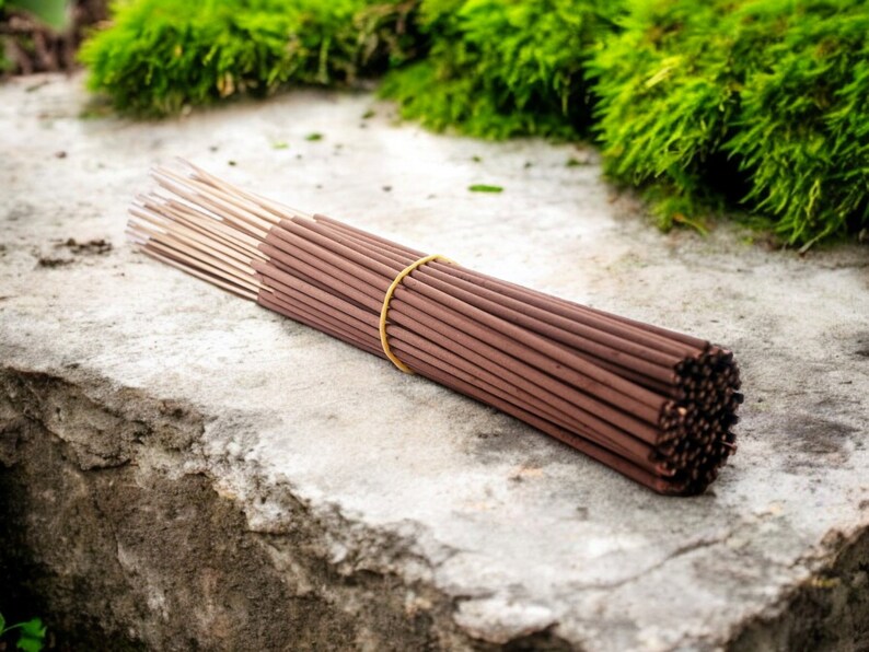May include: A bundle of brown incense sticks tied together with a yellow rubber band. The sticks are arranged in a circular shape and are lying on a gray concrete surface.