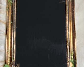 May include: A rectangular gold-colored frame with a black interior. The frame is decorated with green moss and dark crystals at each corner. The frame has an ornate, textured design.