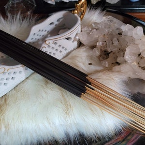 May include: A bundle of black incense sticks with a natural wood base. The sticks are arranged on a white fluffy surface with a white ceramic holder and a cluster of clear quartz crystals.