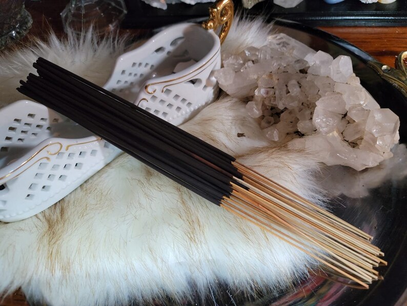 May include: A bundle of black incense sticks with a natural wood base. The sticks are arranged on a white fluffy surface with a white ceramic holder and a cluster of clear quartz crystals.