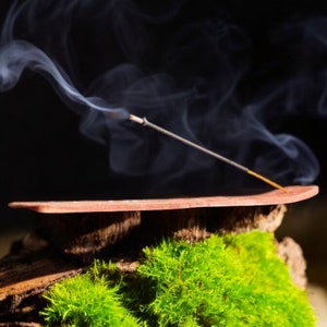 May include: A wooden incense holder with a brown finish, resting on a bed of green moss. A thin incense stick is burning, releasing wispy white smoke.