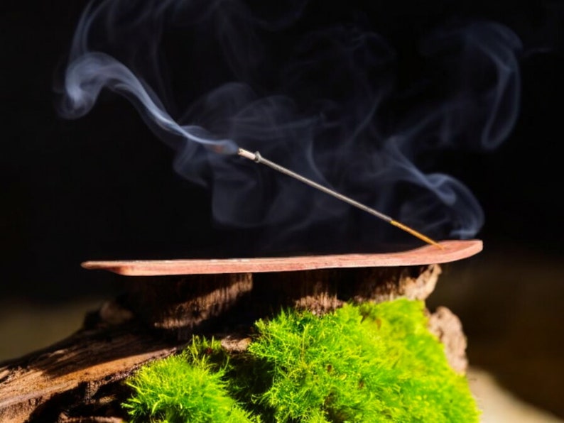 May include: A wooden incense holder with a brown finish, resting on a bed of green moss. A thin incense stick is burning, releasing wispy white smoke.