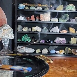 May include: A smoldering incense stick on a silver tray with a decorative design. The stick is burning and releasing smoke. In the background, a shelf displays a collection of colorful crystals and stones.