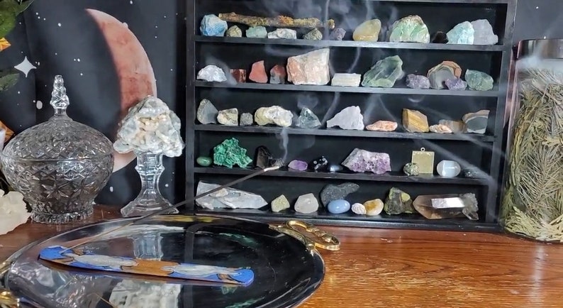 May include: A smoldering incense stick on a silver tray with a decorative design. The stick is burning and releasing smoke. In the background, a shelf displays a collection of colorful crystals and stones.