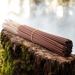 May include: A bundle of brown incense sticks tied together with a yellow rubber band, resting on a mossy tree stump.