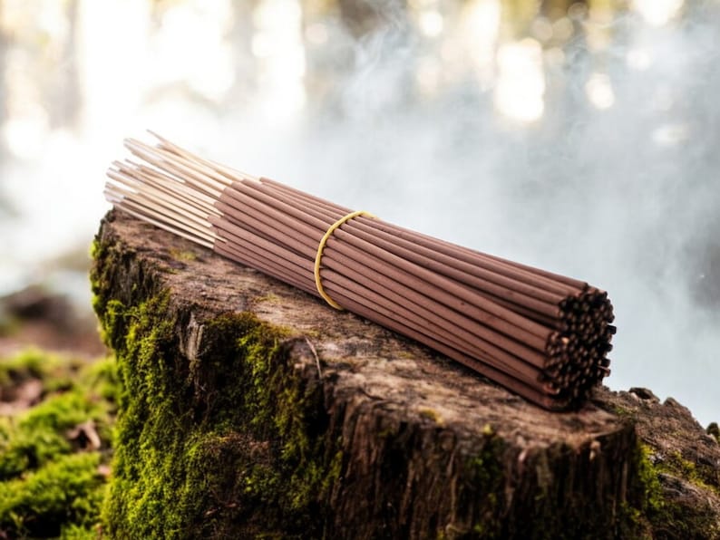 May include: A bundle of brown incense sticks tied together with a yellow rubber band, resting on a mossy tree stump.