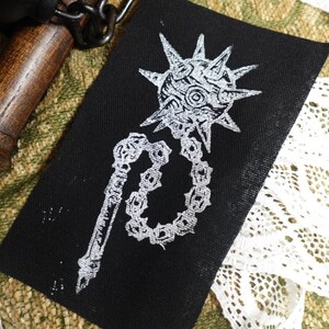May include: Black rectangular patch featuring a white illustration of a spiked mace with a chain. The mace design has a detailed, star-like head and a long handle. The patch is on a textured, patterned background.