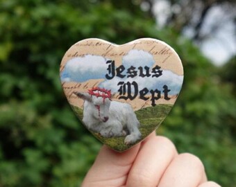 May include: Heart-shaped button with the text "Jesus Wept" in black gothic font. The button features an illustration of a white lamb wearing a crown of thorns, set against a backdrop of clouds and a grassy field.