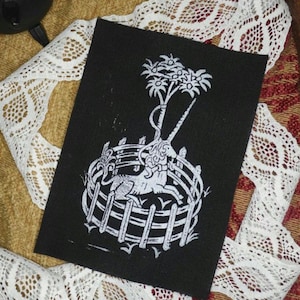 Op de afbeelding: Een zwarte patch met een witte illustratie van een mythisch wezen in een cirkelvormig hek, bekroond met een boom. De rechthoekige patch ligt op een getextureerd oppervlak met witte kanten rand.