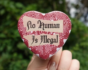 May include: Heart-shaped button with the text "No Human Is Illegal" in a vintage font. The button has a red lace-like border and a cream-colored background for the text. The button is held up against a blurred green background.