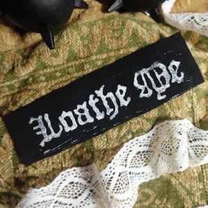 Peut inclure: Un écusson rectangulaire noir avec les mots "Loathe Me" en lettres gothiques blanches. L'écusson est posé sur un tissu vert et beige à motifs, avec une bordure en dentelle blanche. Deux objets noirs en forme de chaudron sont visibles en arrière-plan.
