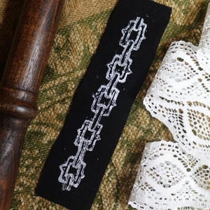 May include: A black fabric tag with a white chain design. The tag is rectangular and features a repeating chain pattern. The image also includes a wooden object, a white lace ribbon, and a textured background.