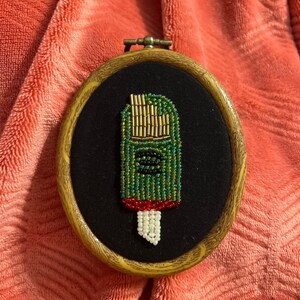 May include: A beaded embroidery of a popsicle on a black background, set in a wooden embroidery hoop. The popsicle design features green, red, white, and gold beads, resembling a frozen treat. The hoop has a metal clasp.