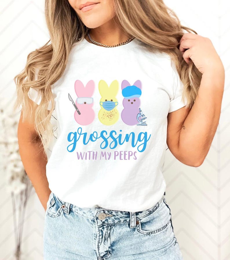 May include: White t-shirt featuring three cartoon Peeps bunnies in pink, yellow, and purple, each with medical-themed accessories. The shirt has the text "grossing with my peeps" in blue script.