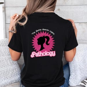 Op de afbeelding: Zwart T-shirt met een roze en zwarte afbeelding. De afbeelding toont een silhouet van een vrouwenhoofd met een paardenstaart in een roze sterpatroon. De tekst luidt "This Barbie dissects organs, Pathology."