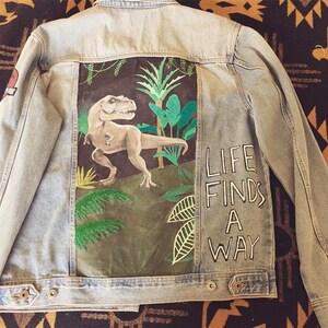 Op de afbeelding: Een lichtblauw denim jack met een handgeschilderd ontwerp op de rug. Het ontwerp toont een groene jungle scène met een Tyrannosaurus Rex dinosaurus en de woorden "Life Finds A Way".