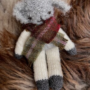 May include: A grey and white knitted sheep doll wearing a red and green tartan scarf. The doll is sitting on a brown and white furry surface.
